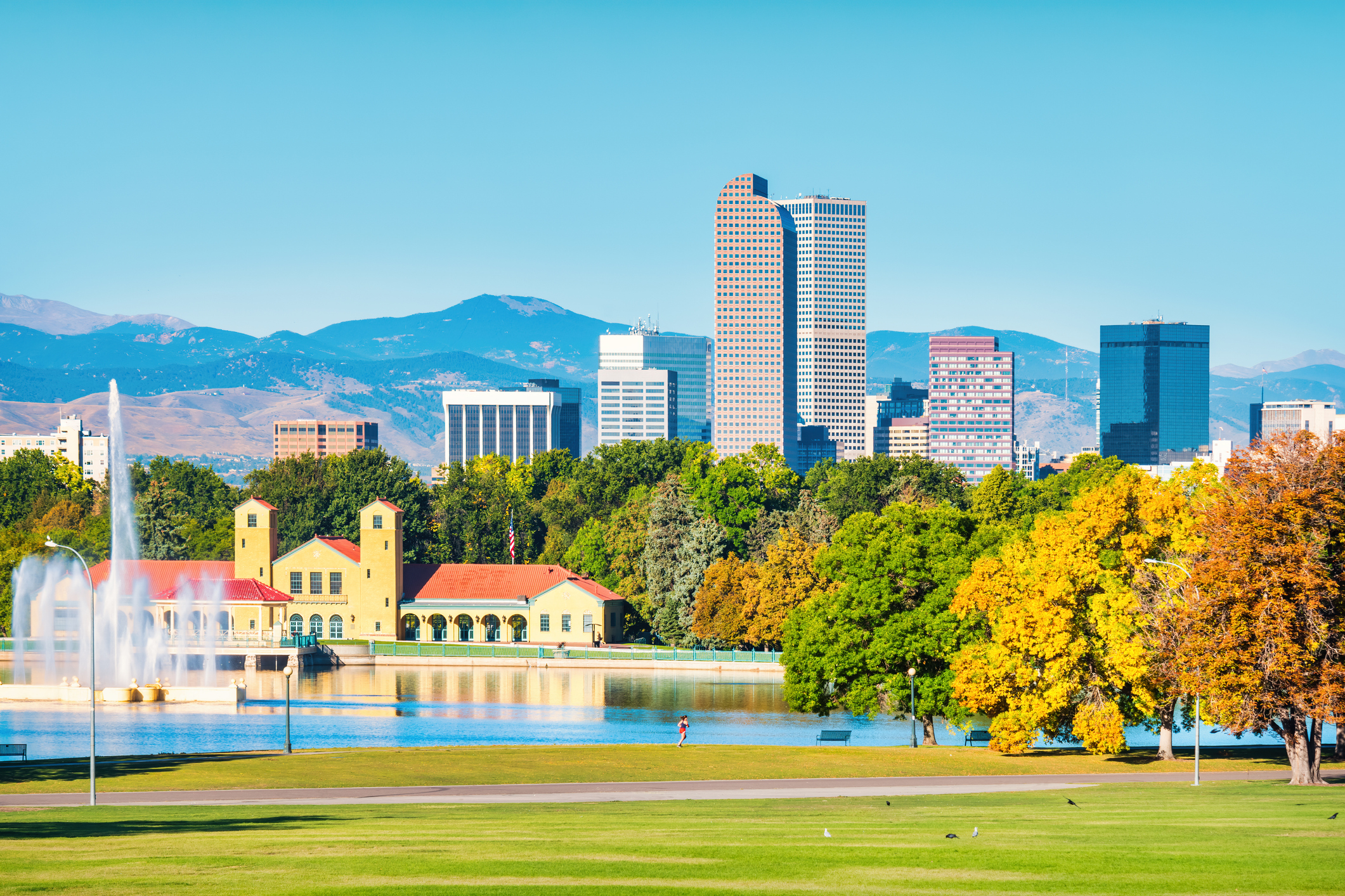 The Denver, Colorado skyline with autumnal trees, blue sky, green fields, a giant fountain and lake, and mountains in the background