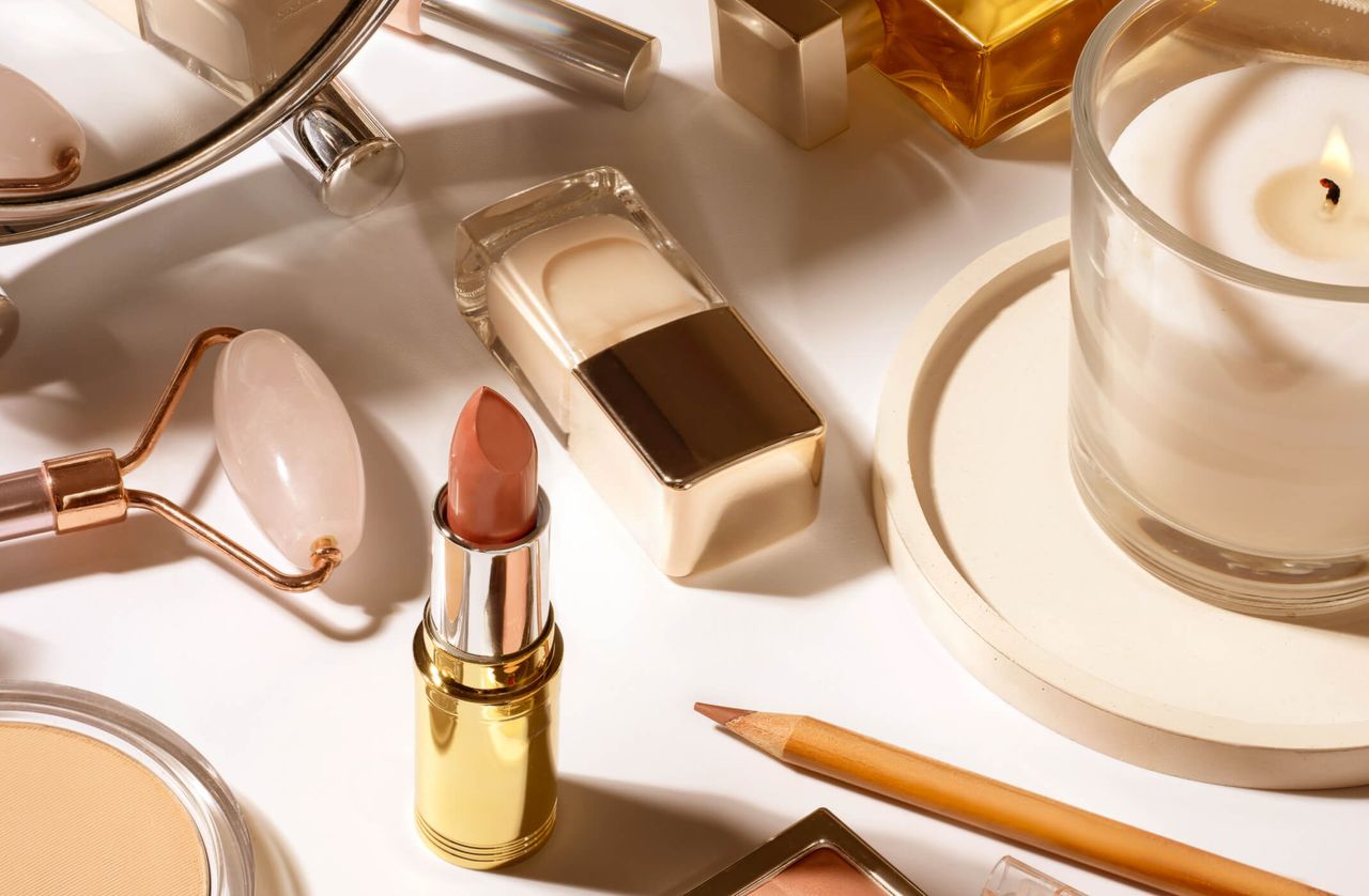 A makeup table with various makeup essentials on like lipstick, a rose quartz roller, and a candle.