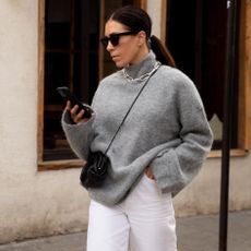 British fashion influencer Jessica Skye poses with a low ponytail, black sunglasses, a gray mock neck turtleneck sweater, silver chunky chain necklace, black crossbody mini bag, and white jeans