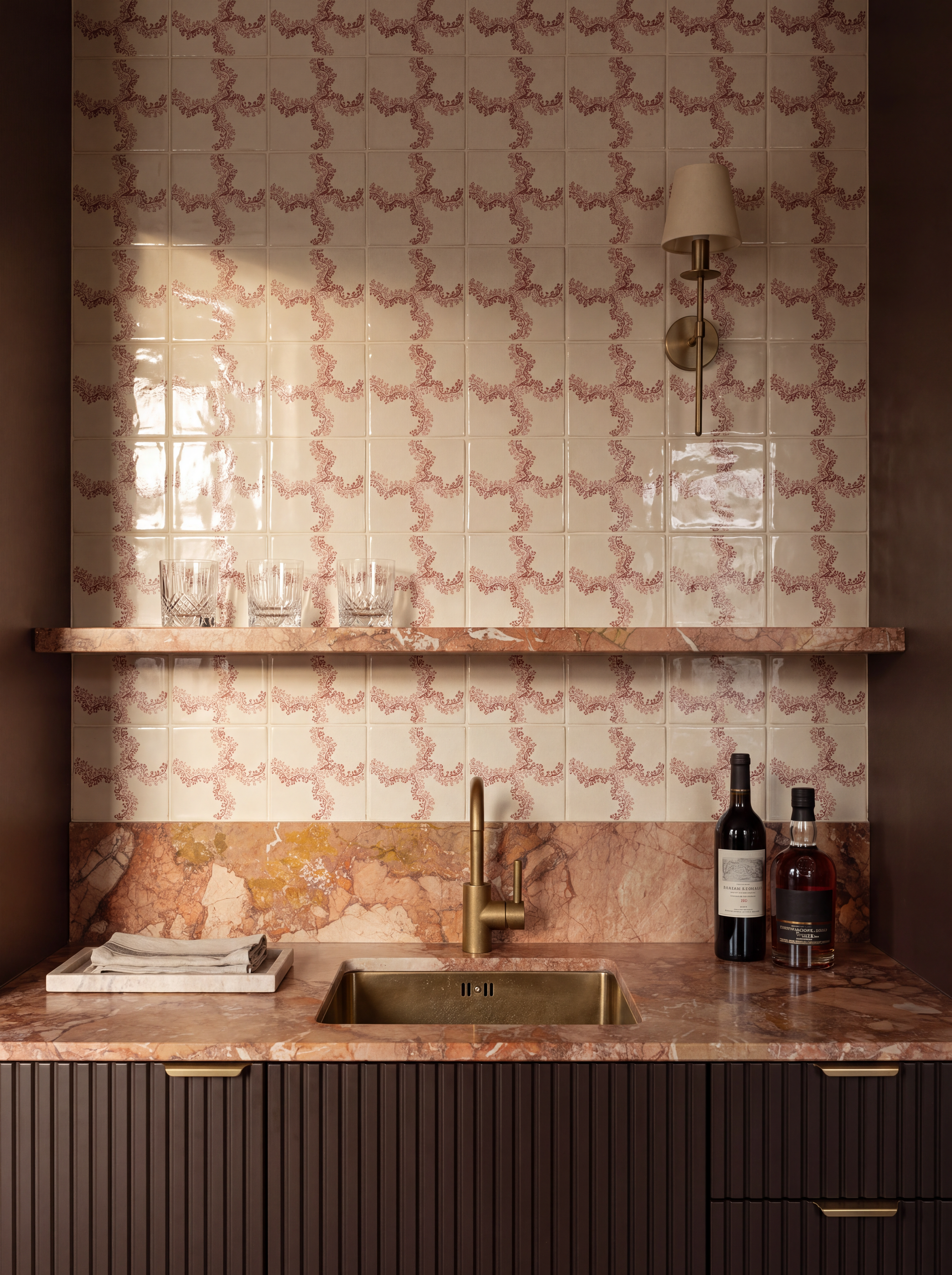 Image of a bar with pink marble countertops, a gold sink, dark wood cabinetry, and pink and white tiles on the wall.