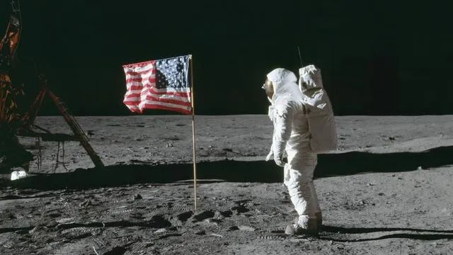 Buzz Aldrin facing the US flag on the moon with the lunar lander off to the left.