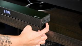 the ultimea nova s50 a black compact soundbar with LED on the front and a wired subwoofer is photographed on a black TV mount connected to a panasonic TV