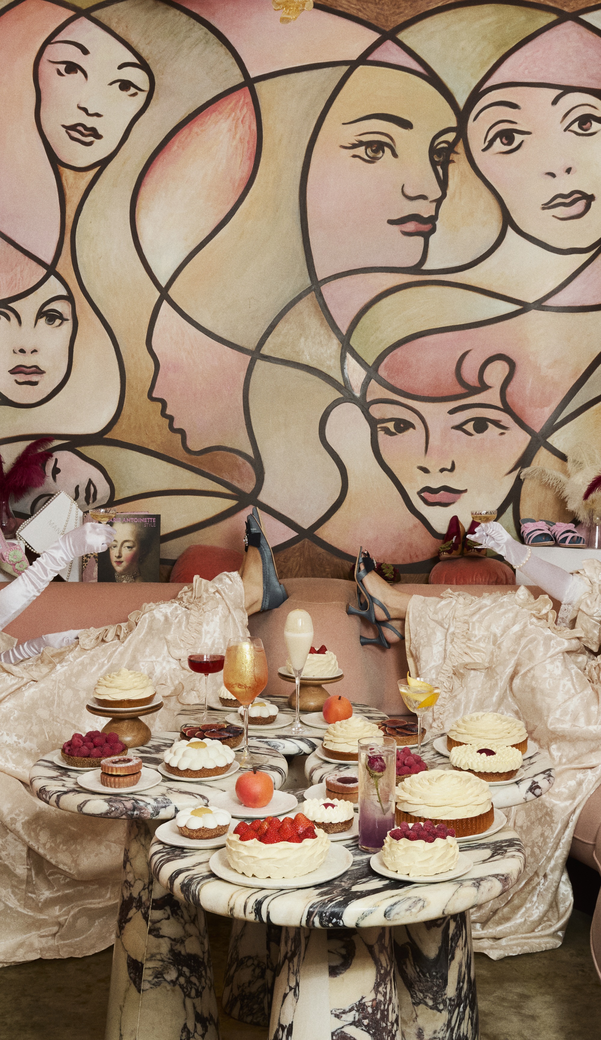 a wall with female faces in a mural with cakes, a cocktails, and two people in dresses with their legs up with shoes on