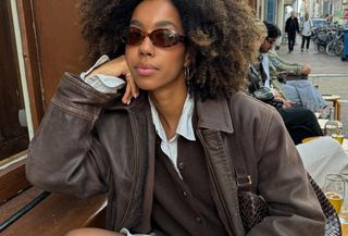 Influencer wearing button down shirt, brown sweater, and brown leather jacket while wearing brown oval sunglasses and sitting on outdoor cafe bench.