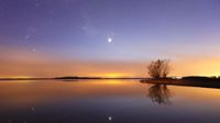 a bright venus shines above a large body of water and is reflected in the water. the sky is hues of blue and orange.