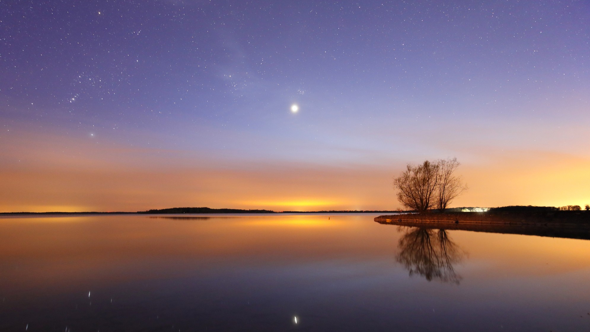 a bright venus shines above a large body of water and is reflected in the water. the sky is hues of blue and orange.