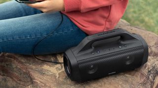 Anker Soundcore Motion Boom Bluetooth speaker on a rock outdoors in the daytime, being used to charge someone's phone.