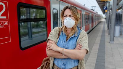 Woman wearing a protective face mask while standing on train platform with her arms crossed, looking annoyed