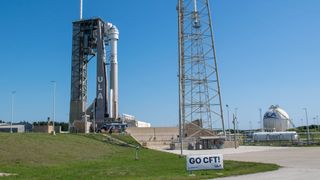a clear blue sky stands behind a rocket launchpad with a tower and rocket standing above a slope of grass.