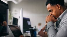 Male software developer with his hands in his face looking frustrated while using AI tools on a laptop computer in an open plan office space.