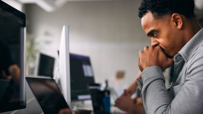Male software developer with his hands in his face looking frustrated while using AI tools on a laptop computer in an open plan office space.