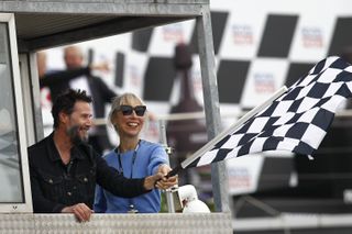 Keanu Reeves in a black denim jacket and Alexandra Grant in a blue sweater and black sunglasses waving a checkered flag together at the MotoGP of Germany at the Sachsenring Circuit on July 07, 2024