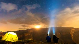 Two people with headlamps looking out at the night sky from atop a rocky cliff at night
