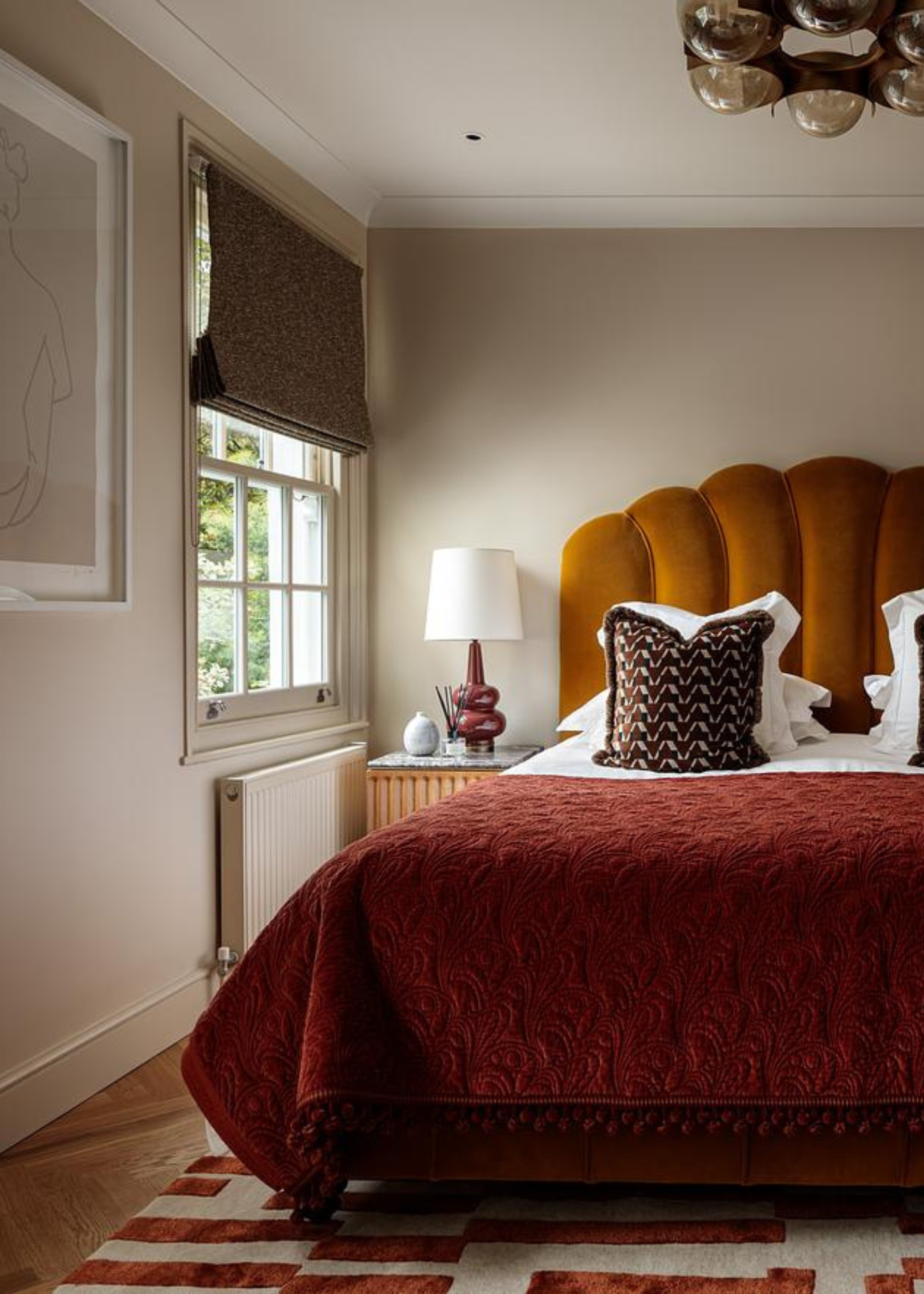 A white bedroom with a large maroon and off-white rug, a bed with a scalloped dramatic headboard and a side table with a lamp by the window