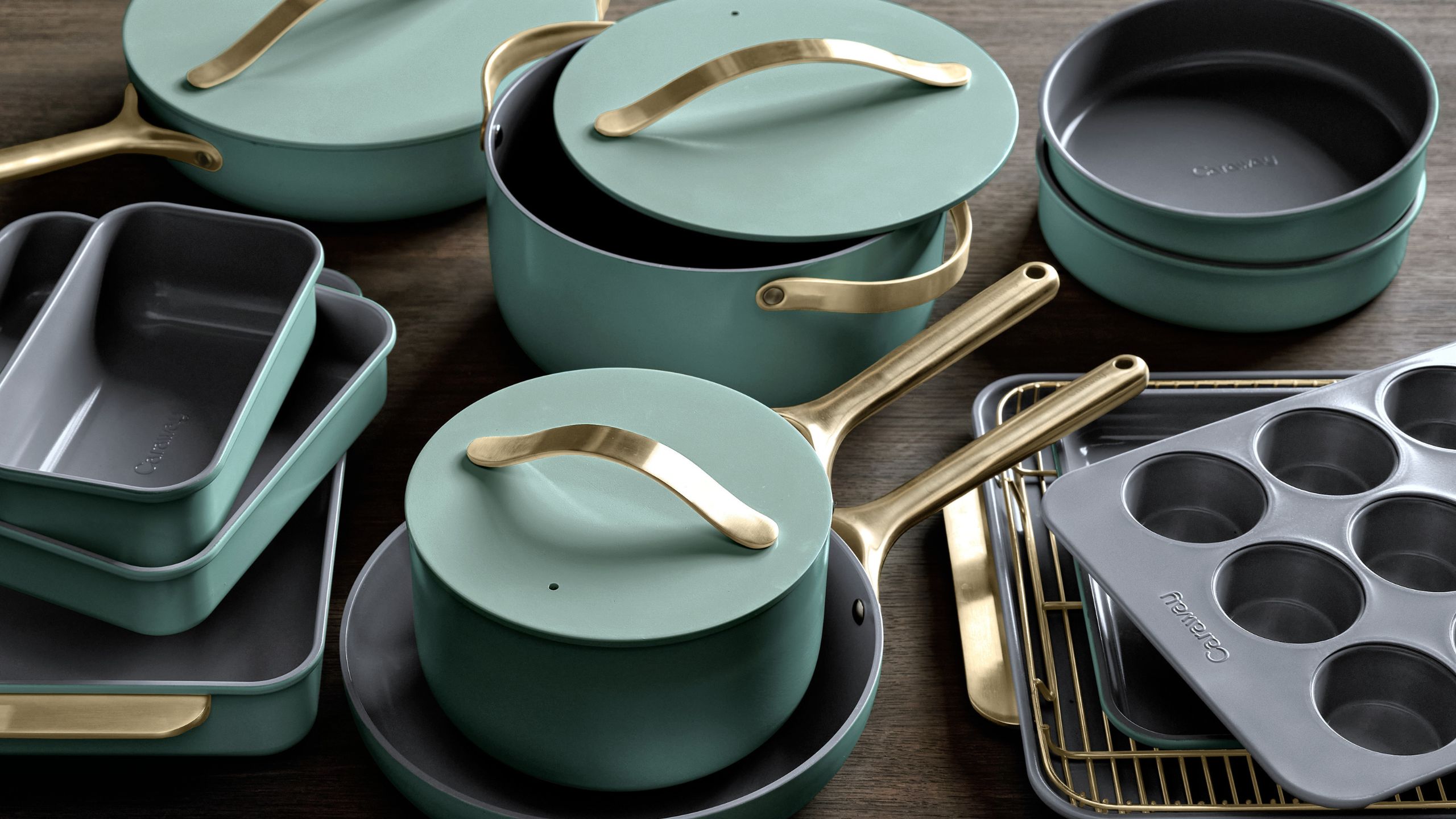 A set of light sage green pans and bakeware with brushed gold handles on a wooden table, viewed from above. 