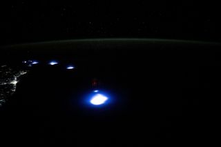 from space, a pitch black earth looms below. faint city lights on the upper left spawn a trail of four blue dots that follow toward the middle, getting larger. A faint layer of green atmosphere is seen at the planets black curve, between it and the black of space, faint with stars.