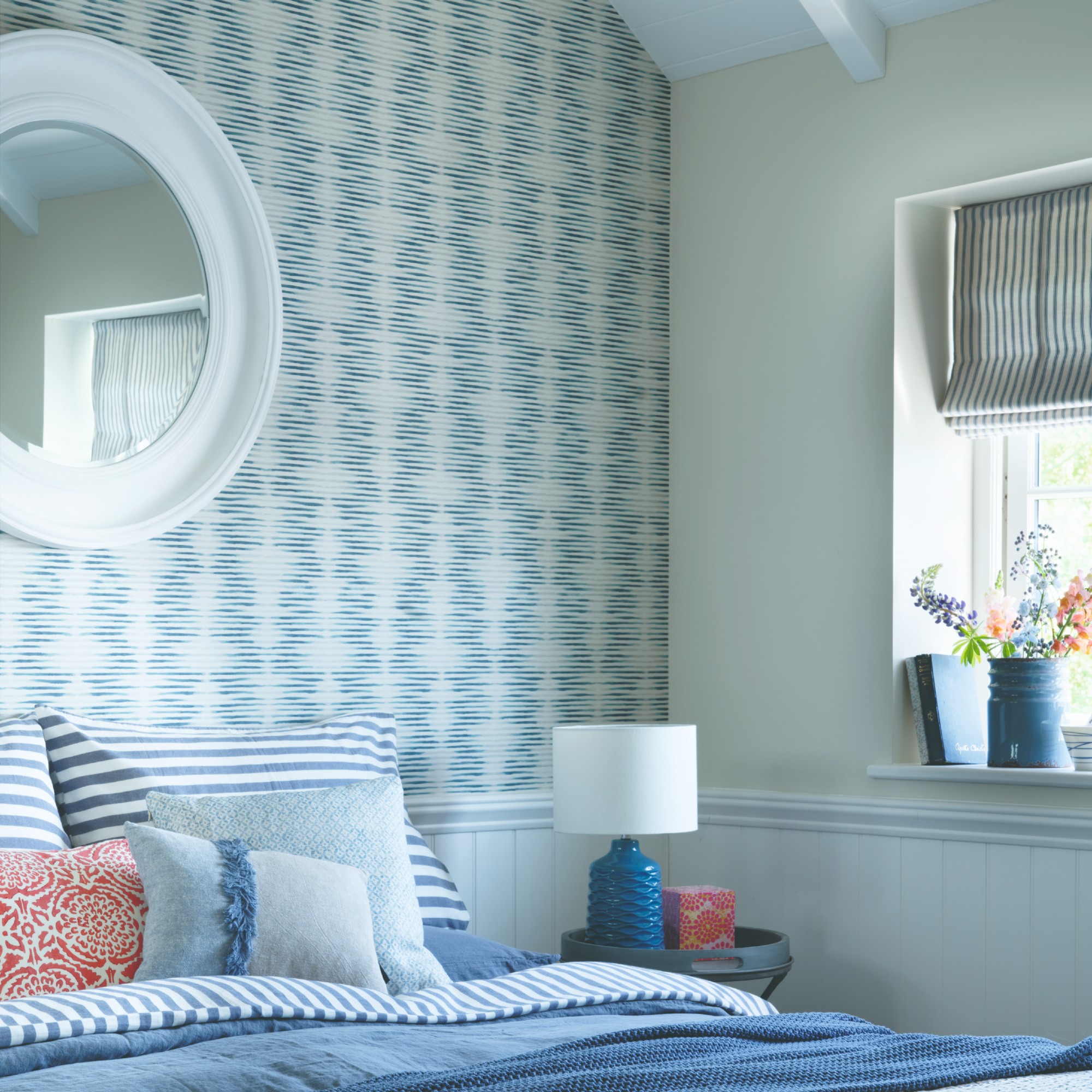 A blue bedroom with the feature wall behind the bed covered in an abstract-print wallpaper and a round mirror