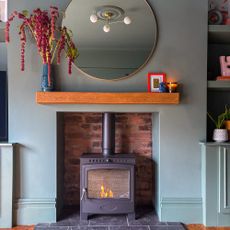 Wooden floating shelf on a grey chimney breast with vintage vase filled with flowers on the left and a modern circular mirror above it. A black wood burner glows in the brick fireplace below