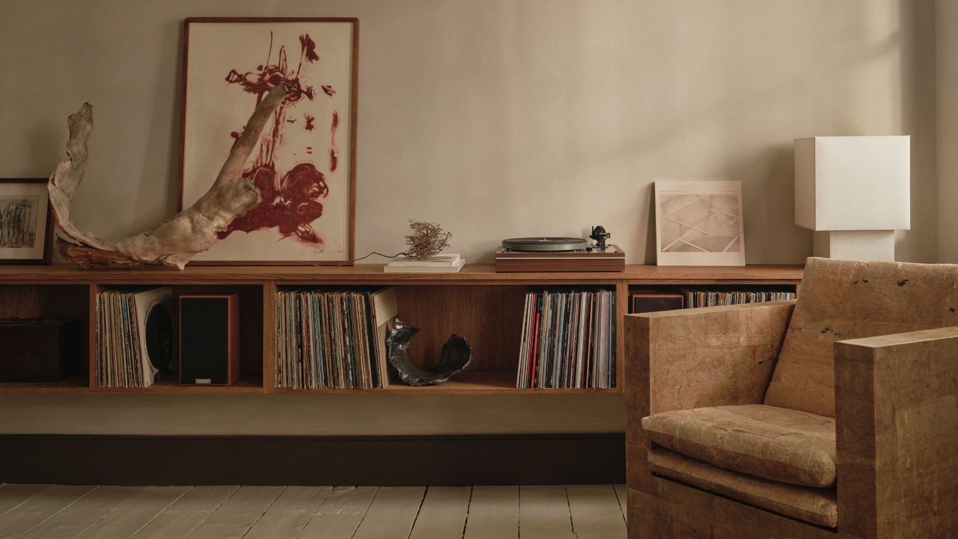 A living room with a textured accent armchair, a floating display of vinyl, a record player, a white table lamp, a leaning painting, and ceramics