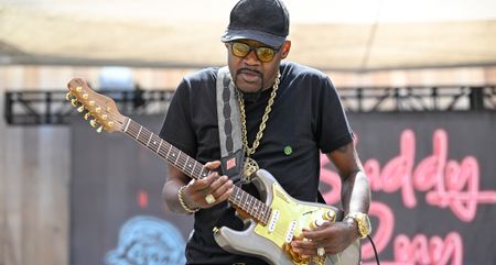 Eric Gales takes a solo on his signature Magneto electric guitar, with its gold pickguard matching his thick gold chain. He wears a ball-cap and tinted glasses.