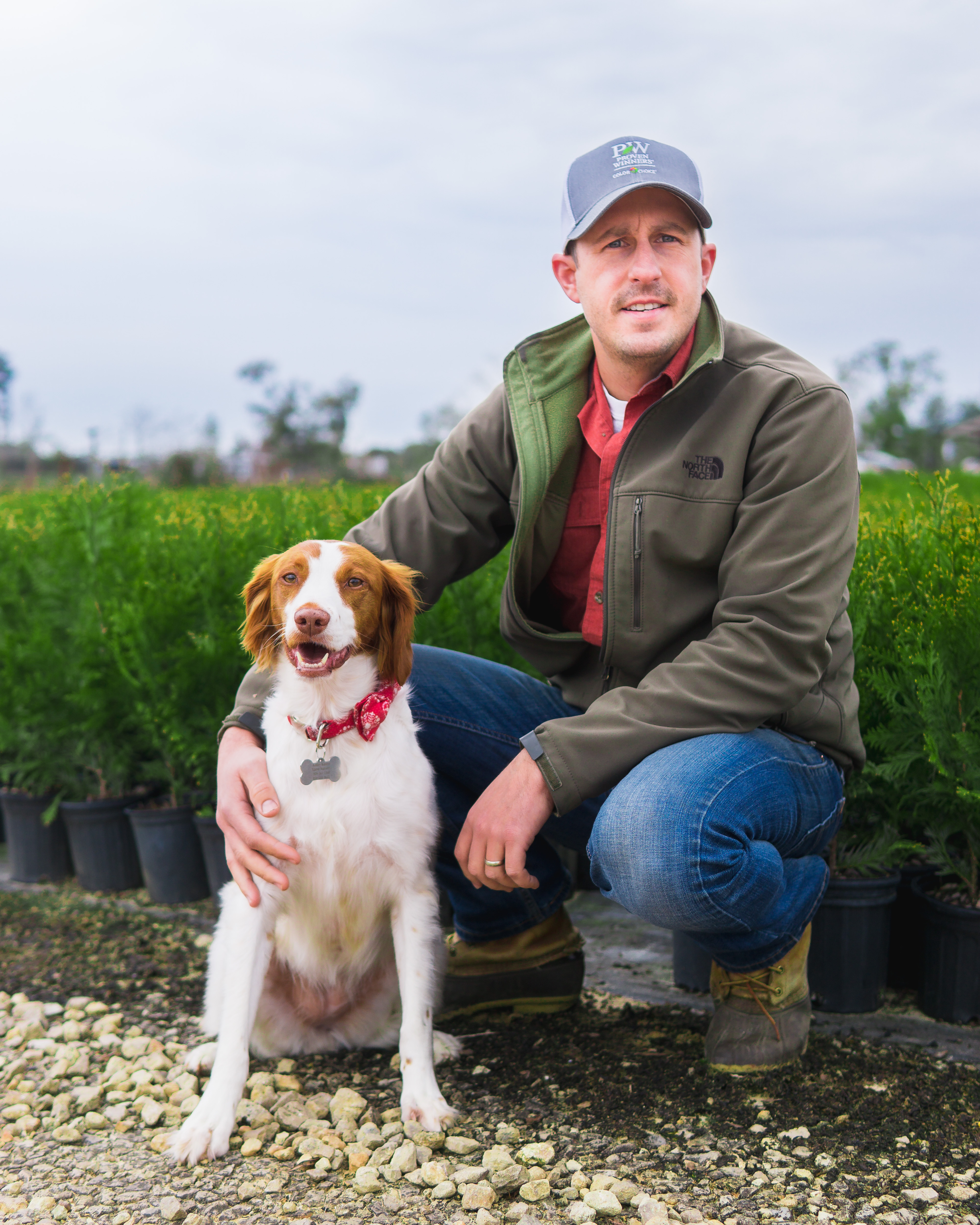 a white man with a baseball cap on crouched down in front of rows of potted plants with a dog