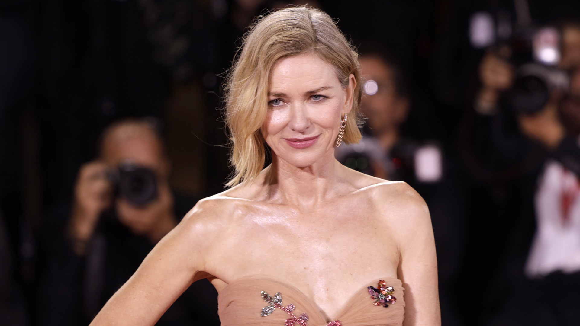 a white woman with short, blonde hair posing on a red carpet wearing a tan, strapless dress with sequins