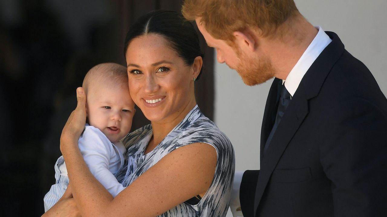 Meghan Markle wearing a blue patterned dress holding baby Prince Archie and smiling while Prince Harry, wearing a suit, looks on