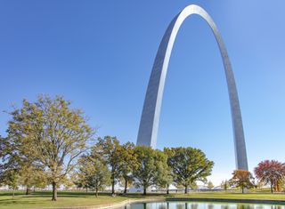 A view of the Gateway Arch in St Louis Missouri from within the park Fall colors adorn the deciduous trees in the foreground and a blue sky completes the scene The arch is inside Gateway Arch National Park and was designed by Eero Saarinen