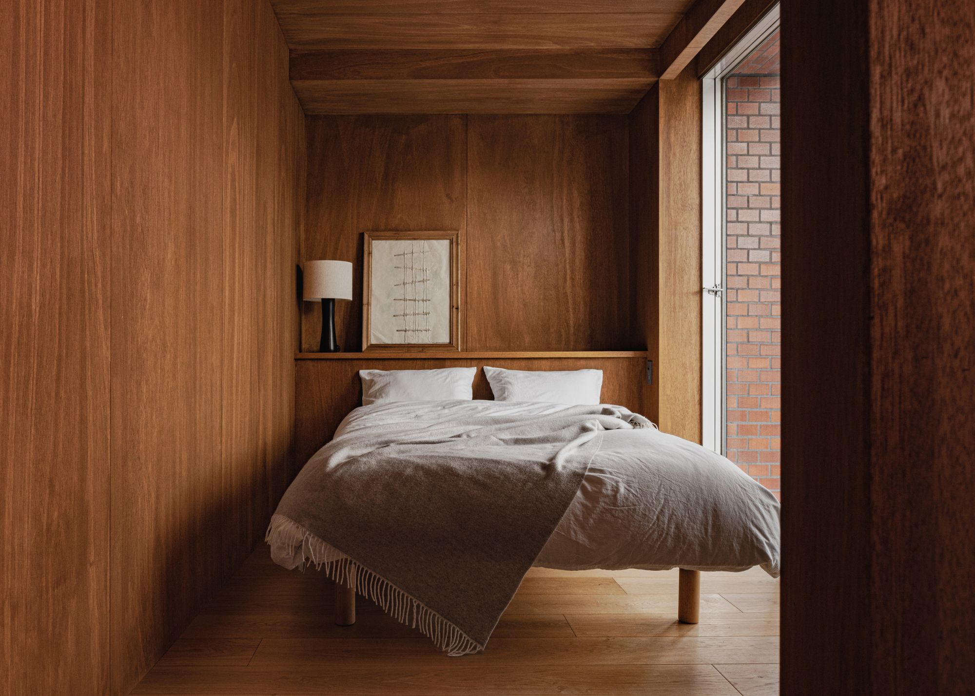 A minimalist Japanese bedroom drenched in wood with just a bed, lamp, and artwork on a ledge above it, next to a floor-to-ceiling sliding glass window/door
