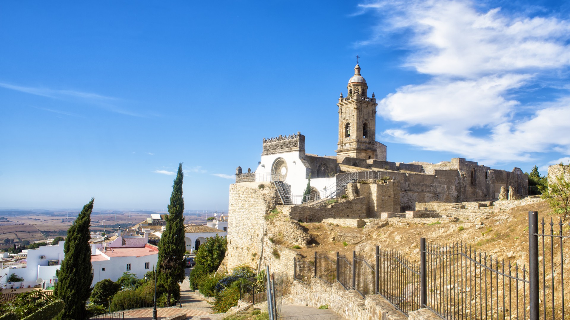 A sunlit stone church and bell tower stand within ancient fortress walls above a town, with cypress trees, dry hills, and a vivid blue sky.