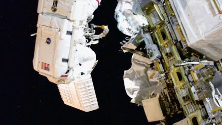 two astronauts in white spacesuits, one holding a piece of equipment, work outside a space station on a spacewalk