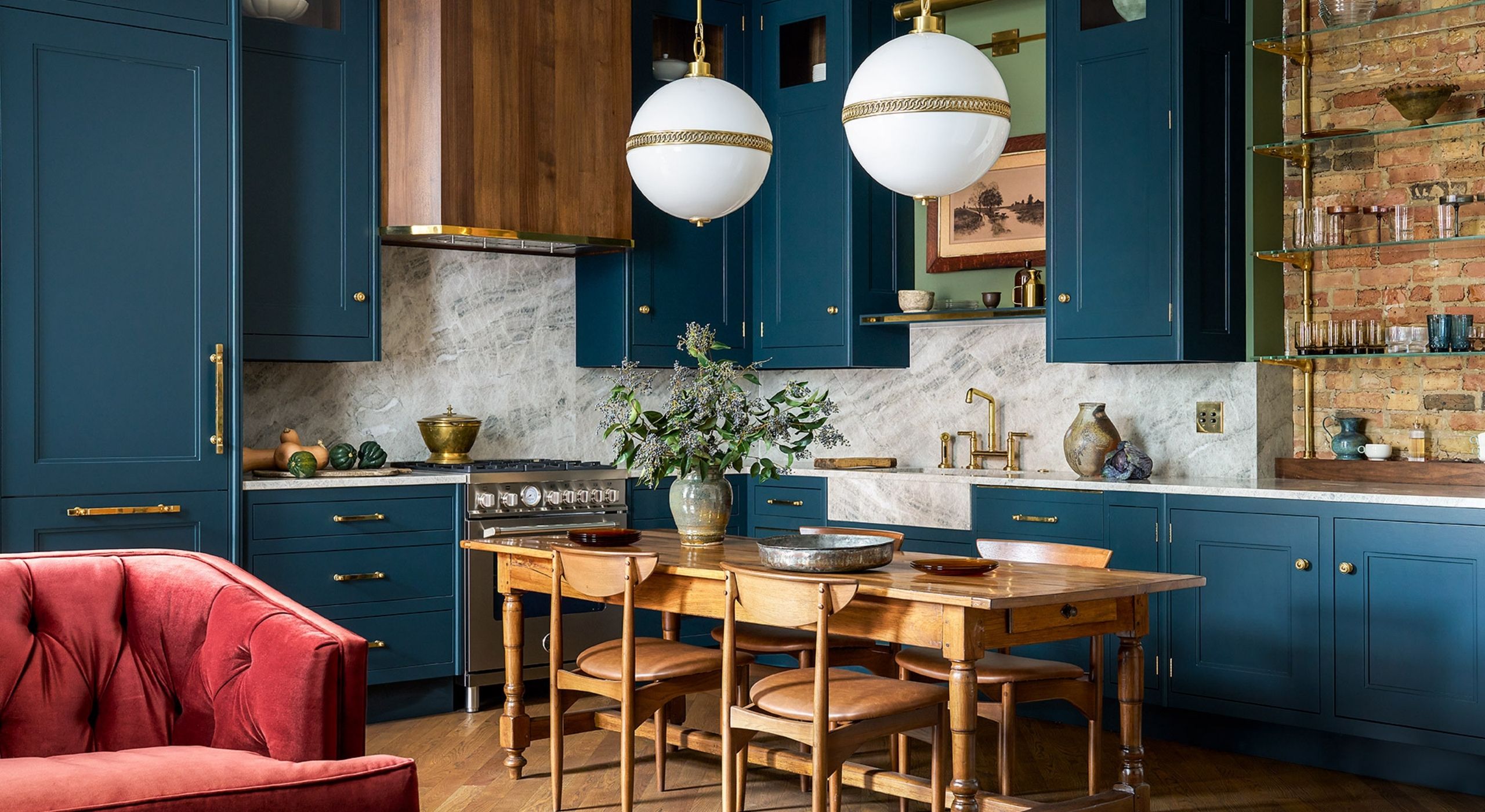 An ocean blue-painted kitchen-cum-living room features a central, long wooden table, spherical art deco pendant lights, a velvet red two-seater sofa, and brick and marble details.