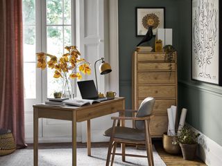 home office with sage green walls, retro style desk and furniture, rug, artwork, autumnal stems in vase on desk, desk lamp