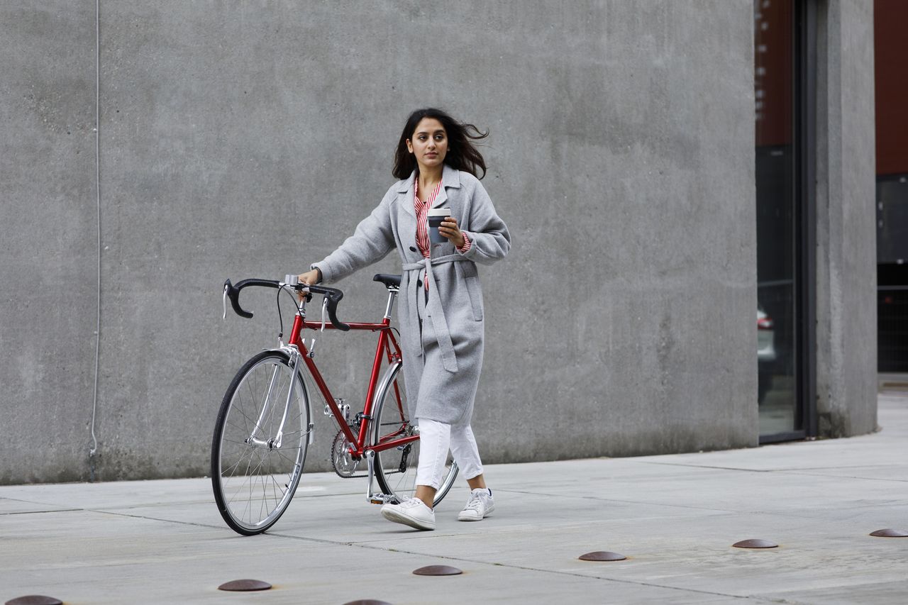 Young woman walking with bicycle, with reusable coffee cup in hand