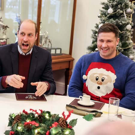 Prince William laughs while wearing a burgundy sweater and a suit jacket and sitting next to a man wearing a blue sweater with a big Santa face on the front