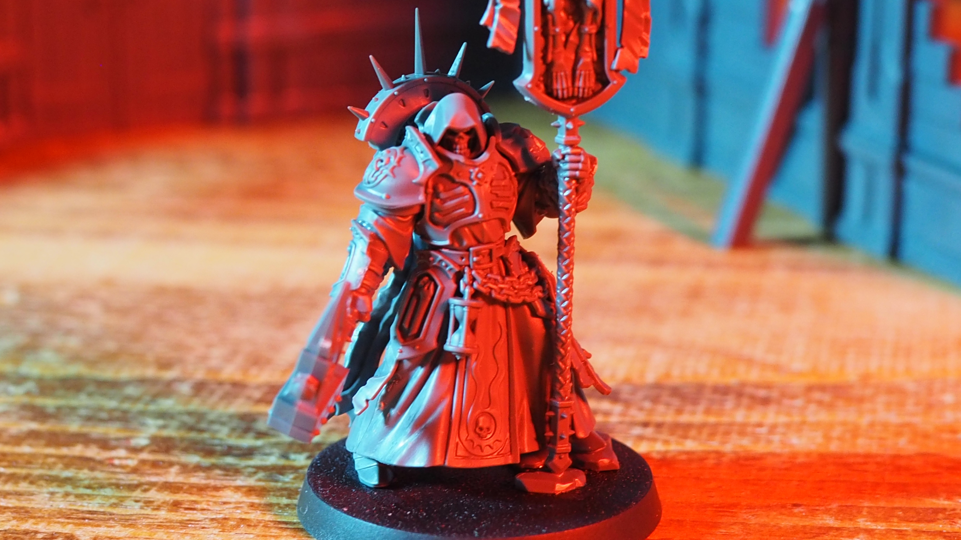 A hooded Lord-Relictor model on a wooden table in front of ruined terrain, all bathed in red light