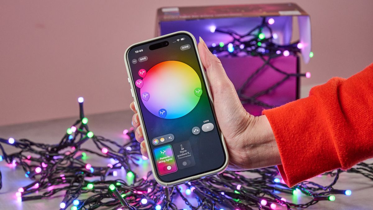 A woman holds a smartphone showing the Philips Hue app. In the background, the Philips Hue Festavia String Lights are glowing in multiple colors while flowing out of their box.