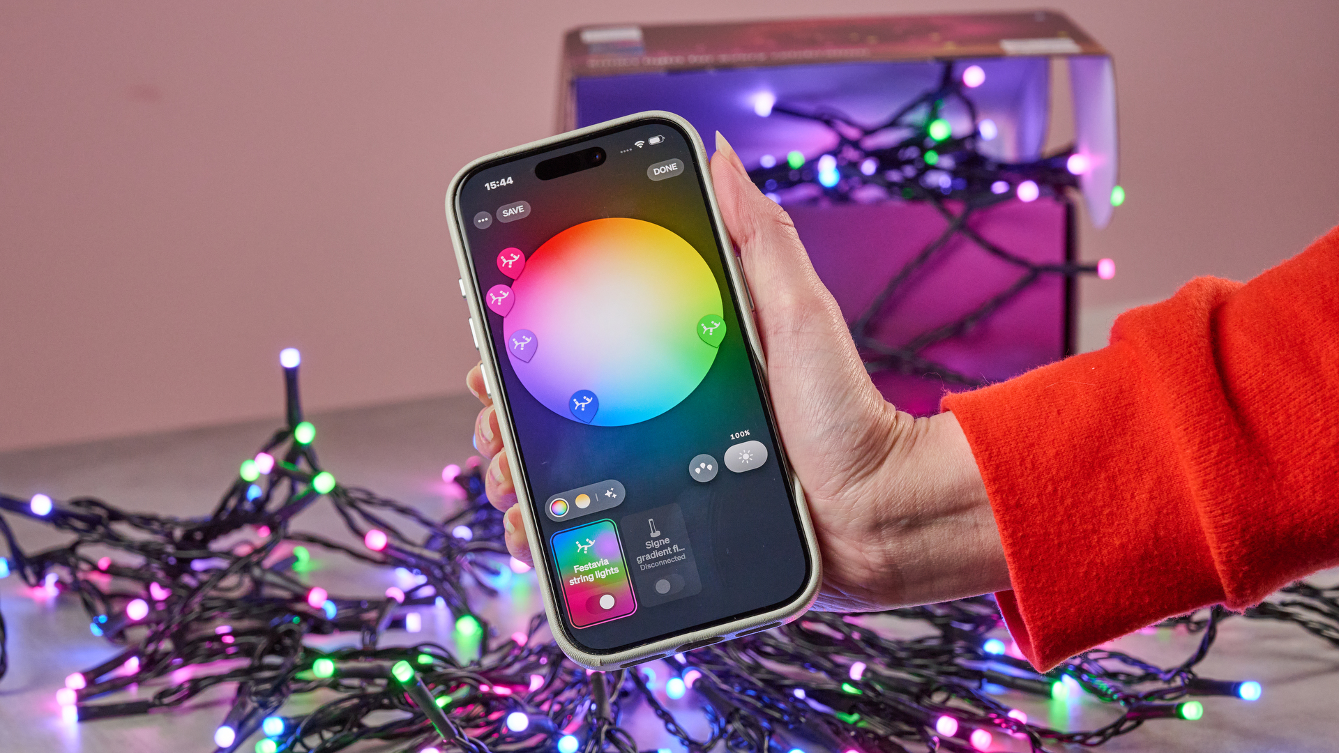 A woman holds a smartphone showing the Philips Hue app. In the background, the Philips Hue Festavia String Lights are glowing in multiple colors while flowing out of their box.