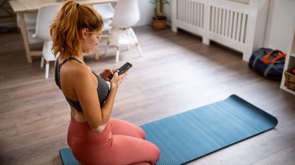 A woman in workout gear, kneeling on an exercise mat, looking at an app on her phone