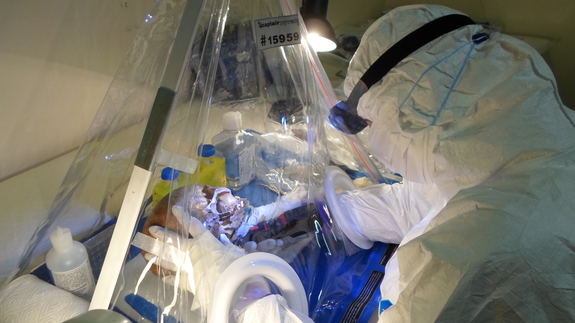 a person in a clean suit handles a human skull through a clear plastic curtain