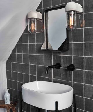 Gray wall tiles with clear bulb sconces, white basin and black faucets