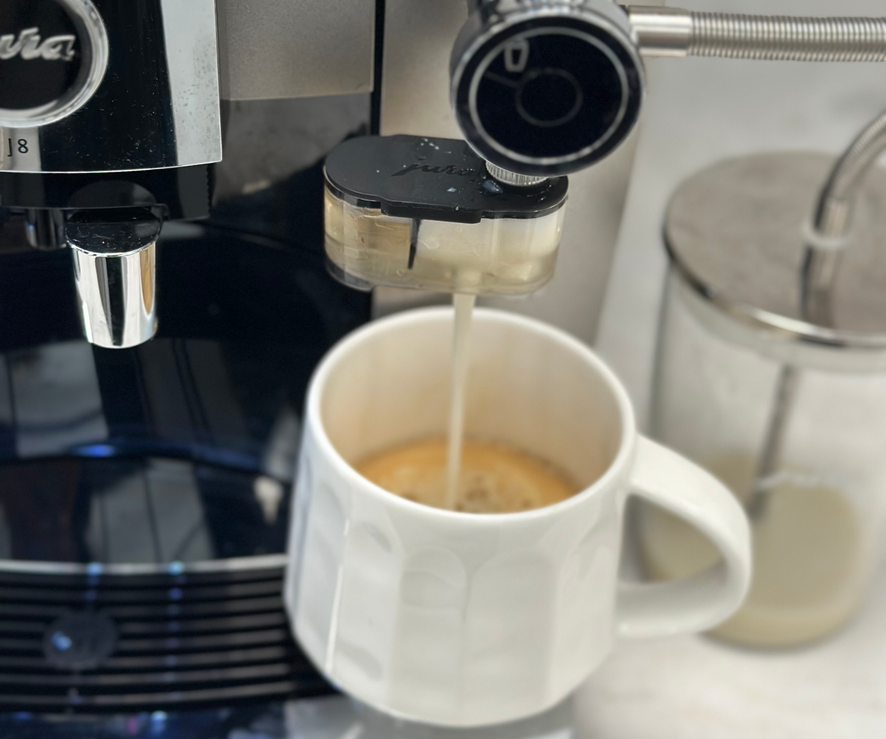 Jura J8 brewing A white mug of coffee under the foam milk spout of a coffee machine.