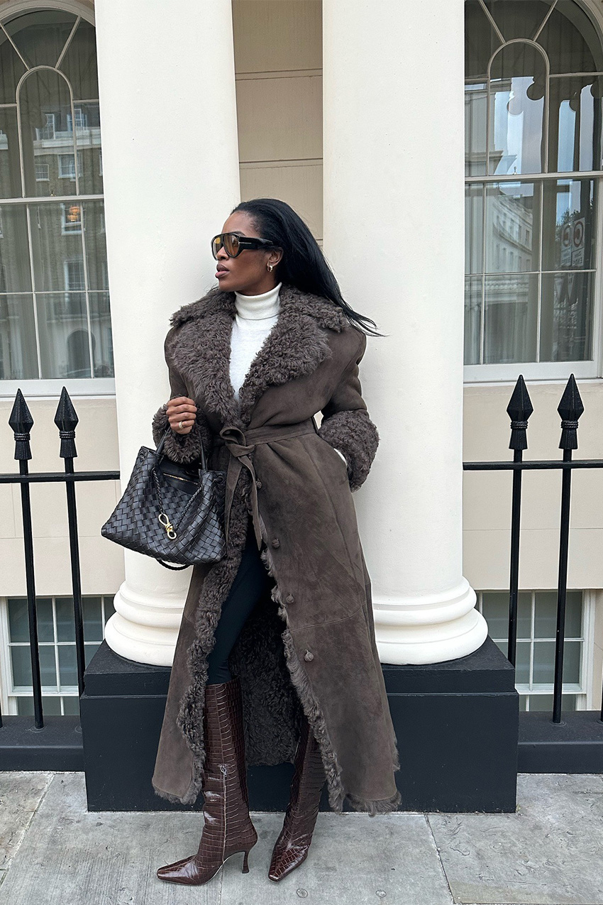The best statement coats are shown in a street style photo of a woman standing on a sidewalk leaning against a white building column wearing oversized two-tone aviator sunglasses with a white turtleneck, black leggings, brown knee-high boots, a brown handbag, and a brown fur and shearling coat
