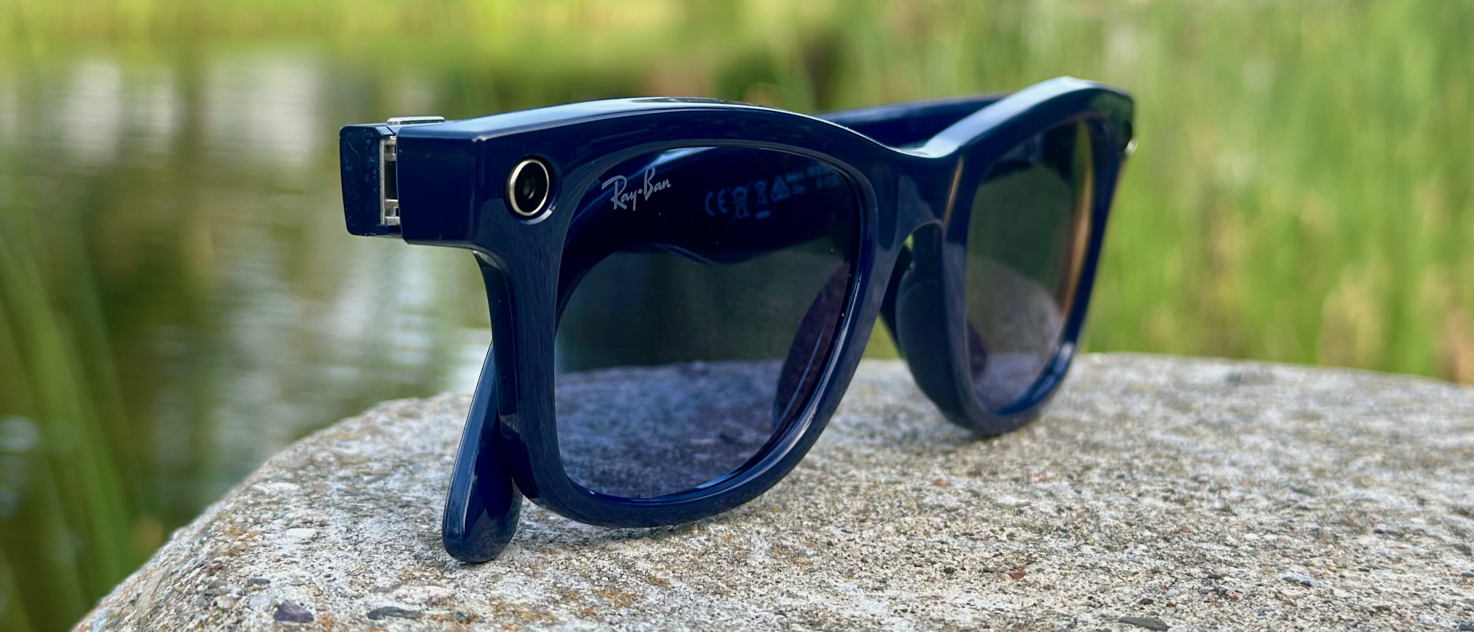 A close-up photo of the right-front side of the Ray-Ban Meta (Gen 2) smart glasses sitting on a rock in front of a pond with reeds, the perspective close to the right camera cut-out and right lens.