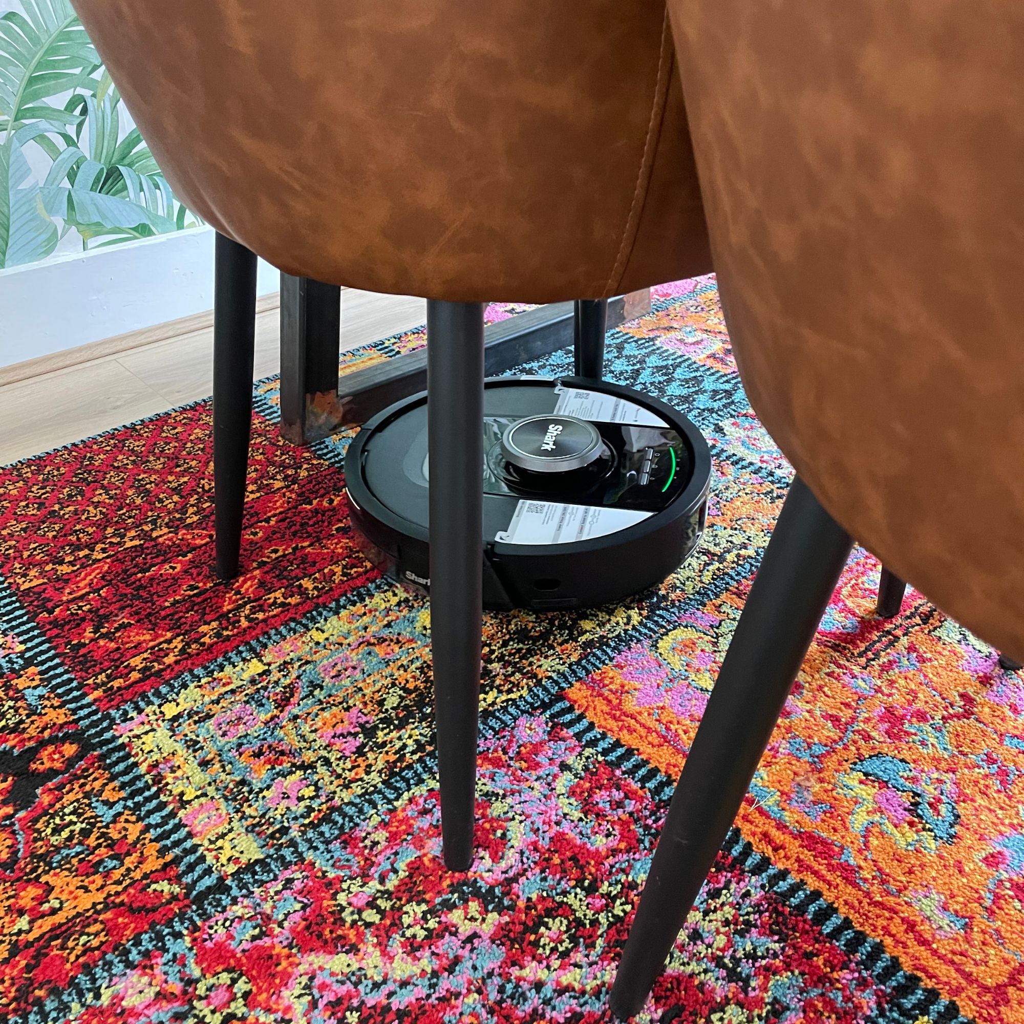 Shark PowerDetect NeverTouch Robot Vacuum cleaning in between the legs of dining room table, on top of colourful rug