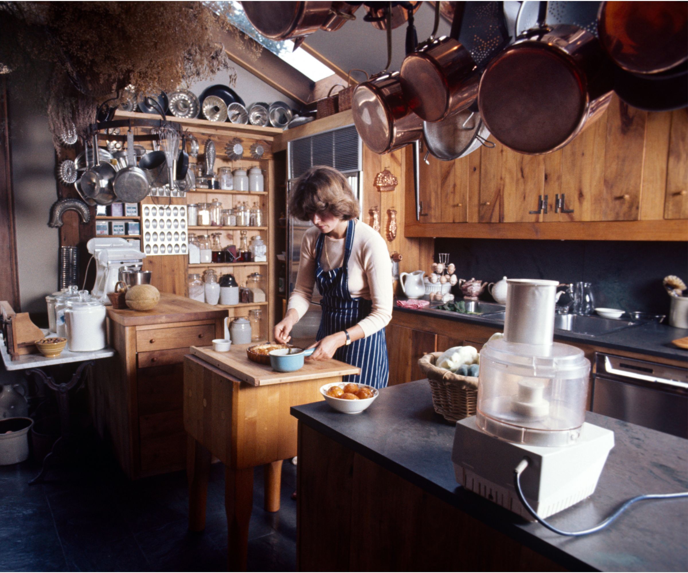 Martha Stewart in her farmhouse kitchen with wood kitchen cabinets, copper pans hanging on rail, pantry with jars and copper molds