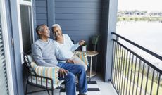 A senior African-American couple sitting side by side on their porch or balcony, relaxing, and holding hands, conversing and laughing together.