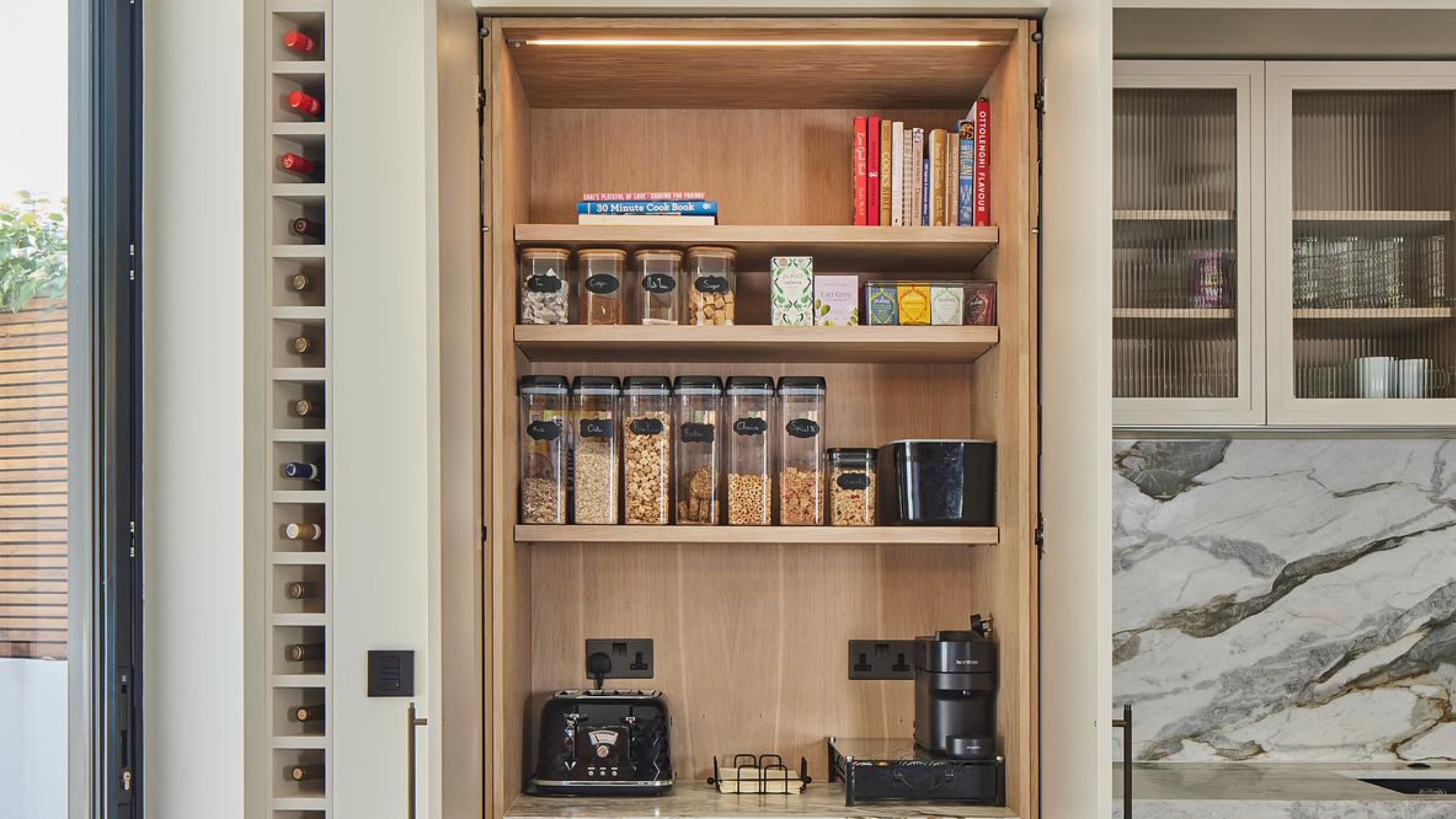 An open pantry unit with jars and a hidden nook for coffee and tea. On the top shelf of the pantry are some books. Outside of the pantry is a view of a wine rack as well as glass fronted cabinets 