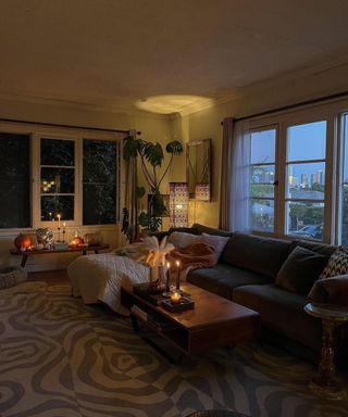 A small living room with multiple light sources including side lamp, table candles and uplighting, a gray couch, a coffee table, and a large window
