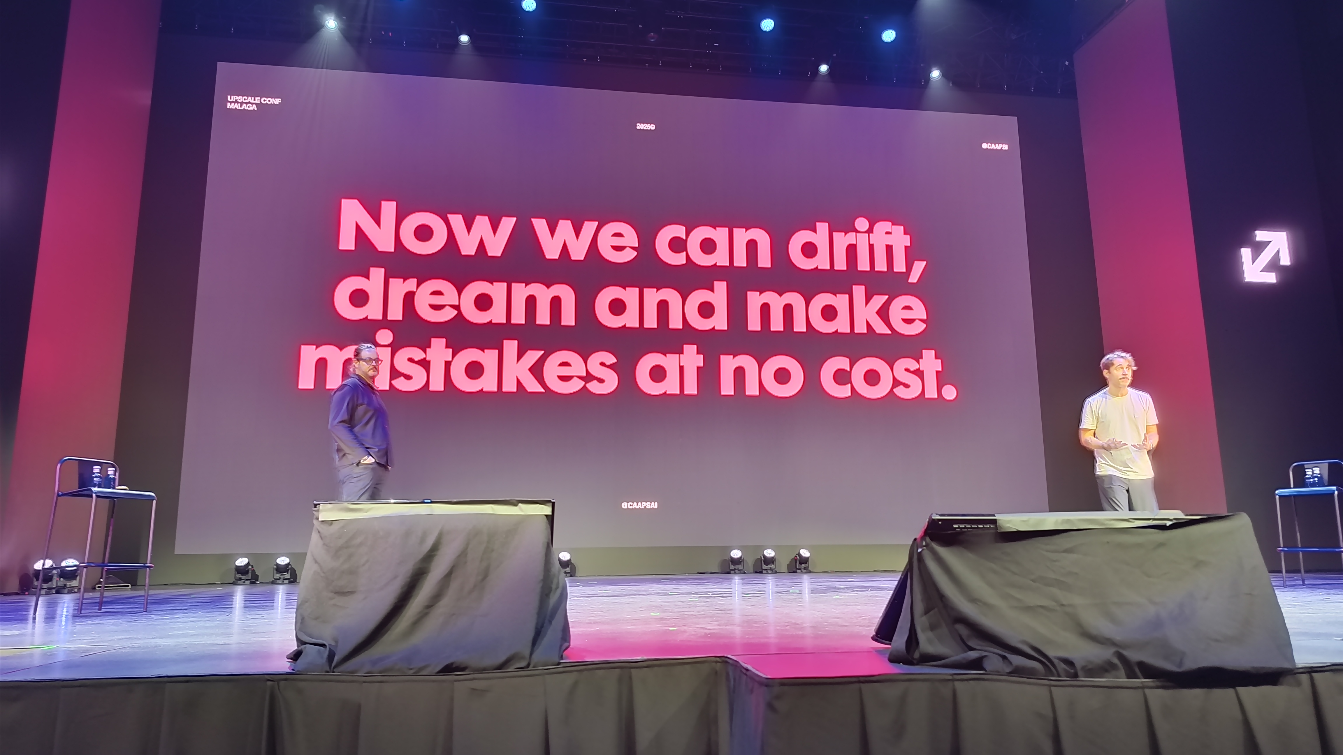 A wide-angle photo of two men standing on a stage during the Upscale Conference in Malaga, with a large screen behind them displaying the red text: &quot;Now we can drift, dream and make mistakes at no cost.&quot;